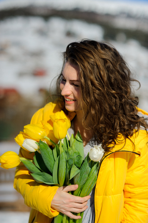 Charming young woman with a bouquet of yellow and white tulips. Curly hair, nice smile. Bright yellow coat blends with the flowers. Spring mood.の写真素材