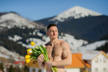 Young man with a naked torso holds a bouquet of tulips. Sunny day. Blue sky. Guy poses against the background of snow-covered mountain tops.の写真素材