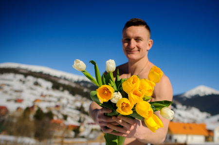 Young man with a naked torso holds a bouquet of tulips. Sunny day. Blue sky. Guy poses against the background of snow-covered mountain tops. He smiles.の写真素材
