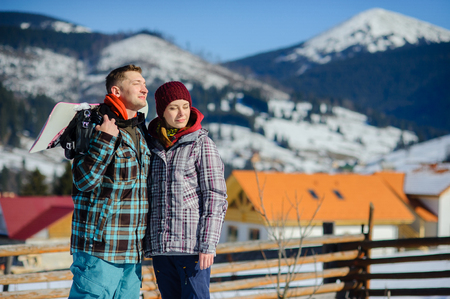 Young couple against the background of a mountain landscape. The guy and the girl admire fine landscapes. Serene winter day, blue sky, snow-covered mountain tops. Behind shoulders of the young man a snowboard.の写真素材