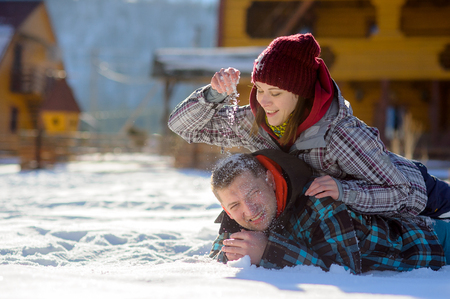 Young couple lies on the snow. The girl climbed onto the back of her boyfriend and sprinkled it with snow. Between them comic fight. Sunny winter day. Good mood. Young people smileの写真素材