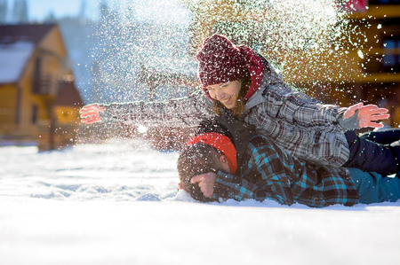 Young couple lies on the snow. The girl climbed onto the back of her boyfriend and sprinkled it with snow. Between them comic fight. Sunny winter day. Good mood. Young people smileの写真素材