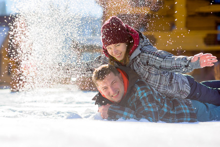Young couple lies on the snow. The girl climbed onto the back of her boyfriend and sprinkled it with snow. Between them comic fight. Sunny winter day. Good mood. Young people smileの写真素材