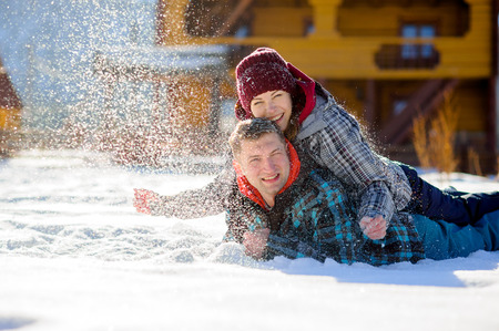 Young couple lies on the snow. The girl climbed onto the back of her boyfriend and sprinkled it with snow. Between them comic fight. Sunny winter day. Good mood. Young people smileの写真素材