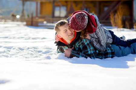 Young couple lies on the snow. Girl climbed onto the back of her boyfriend. She kisses the guy on the cheek. Sunny winter day.の写真素材