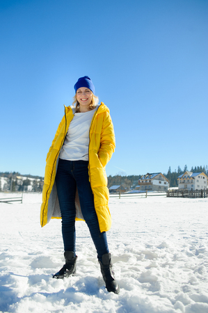 Beautiful young woman in a bright yellow down-padded coat poses for the camera. Serene winter day. Brightly the sun shines. Woman has a great mood. Against the background of a picturesque winter landscapeの写真素材