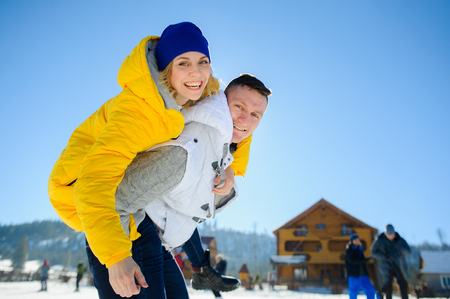 Young man holding his girlfriend on his shoulders. Girl embraces guy for neck. Young people look in the camera and cheerfully smile. Against the background of mountains and blue sky.の写真素材
