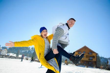 Guy and a girl are having fun in the courtyard of a cottage in a ski resort. A young man carries a girl on her back. She is laughing. Good winter day. Sun, sky, mountains.の写真素材