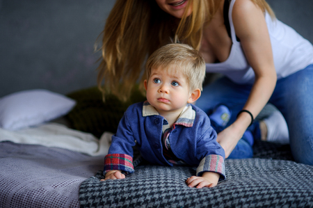 Amusing little boy lies on a big bed. Near him his young mother. She tenderly and carefully concerns the kid. Boy has a cute serious face, beautiful big eyes.の写真素材