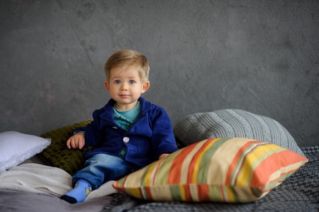 Little boy sits on a bed. Near him big pillows. Child has a touching and pleasant appearance. Chubby cheeks, big blue eyes, long eyelashes, a thick fair hair and bright full lips. Kid has a serious look.の写真素材