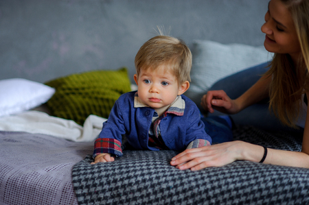 Amusing little boy lies on a big bed. Near him his young mother. She tenderly and carefully concerns the kid. Boy has a cute serious face, beautiful big eyes.の写真素材