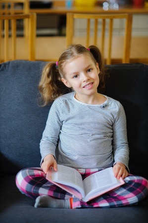 Cute girl of younger school age reads the book. She sits having crossed legs on a soft sofa. The open book lies at the child on a lap. Girl has distracted from reading and looks with a smile in the camera.の写真素材
