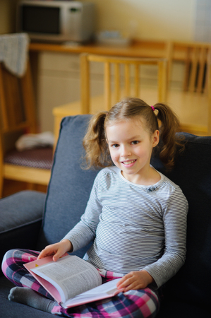 Cute girl of younger school age reads the book. She sits having crossed legs on a soft sofa. The open book lies at the child on a lap. Girl has distracted from reading and looks with a smile in the camera.の写真素材
