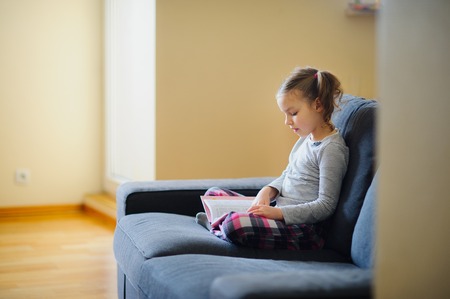The nice girl of younger school age reads the book. She sits having crossed legs on a soft sofa. The girl is keen on reading.の写真素材