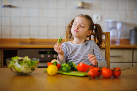 Little girl eating broccoli. She sits at the kitchen table. In front of her a variety of vegetables and herbs. On the table there is a salad bowl with the sliced vegetables. Tasty and healthy.の写真素材