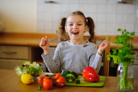 Little girl eating broccoli. She sits at the kitchen table. In front of her a variety of vegetables and herbs. On the table there is a salad bowl with the sliced vegetables. Tasty and healthy.の写真素材
