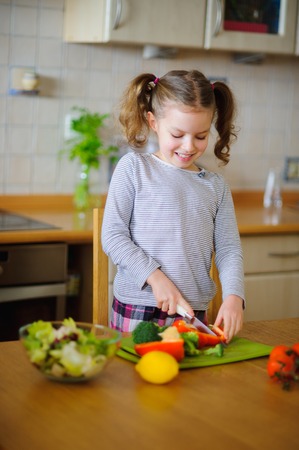 Little cook cooks salad. On a table various vegetables. Lovely girl of younger school age cuts vegetables and greens on a chopping board. Mother's assistant diligently prepares.の写真素材
