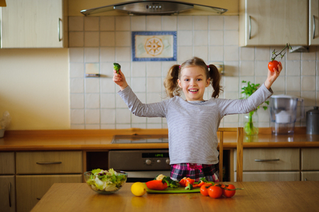 Little girl sits at a table in kitchen. She cut vegetables for salad. Omnifarious veggies and greens are on a kitchen table. The girl likes taste of natural products. Healthy food since the childhood.の写真素材