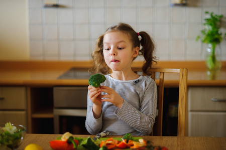 Little girl eating broccoli. She sits at the kitchen table. In front of her a variety of vegetables and herbs. On the table there is a salad bowl with the sliced vegetables. Tasty and healthy.の写真素材