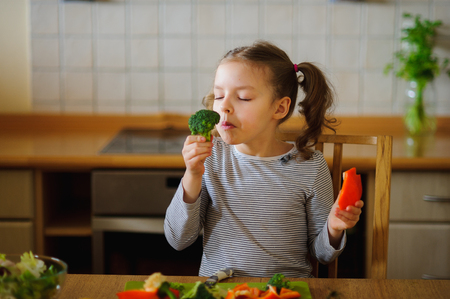 Girl of 8-9 years sits at a kitchen table. Before her various vegetables. In hands at child broccoli and red paprika. The girl is going to eat them. She has closed eyes from pleasure.の写真素材