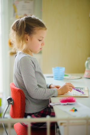 The schoolgirl of elementary school does homework. The girl sits at a school desk and with concentration reads. On table school supplies and different personal objects. To the child it is comfortable.の写真素材