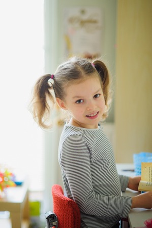 The little schoolgirl performs homework. The girl sits at a desk half-turned to the camera and smiles. On a table school supplies and different personal objects. To the child it is comfortable.の写真素材