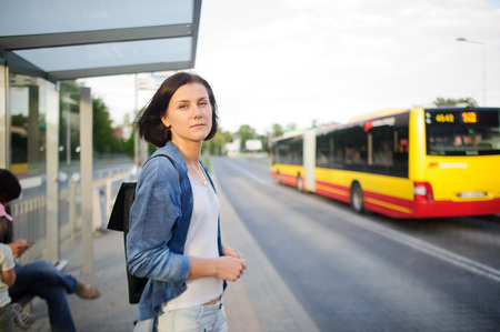 The young woman the brunette stands on the bus stop. She expects the bus. The woman is dressed in sports style. She high and slim. Behind shoulders at her a backpack.の写真素材