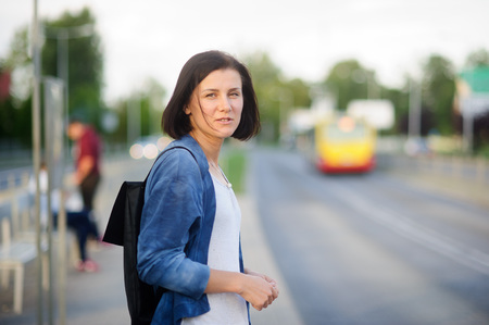 The young woman the brunette stands on the bus stop. She expects the bus. The woman is dressed in sports style. She high and slim. Behind shoulders at her a backpack.の写真素材