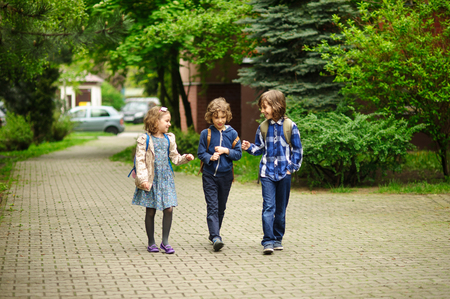 Three little friends go to school. Behind shoulders at children schoolbags. Warm spring morning. To little school students cheerfully together.の写真素材