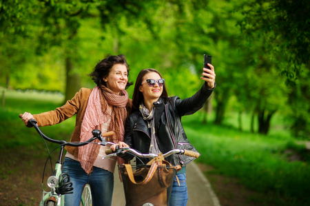 Two nice young women make selfies during bicycle walk in the park. Fine weather. Girlfriends have an excellent mood. They smile.の写真素材