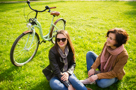 Two young women have a rest on a green lawn. The bicycle stands nearby. Cute girlfriends with enthusiasm about something talk. Fine spring day.の写真素材