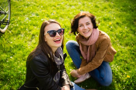 Two young women have a rest on a green lawn. Cute girlfriends joyfully smile. Fine spring day.の写真素材