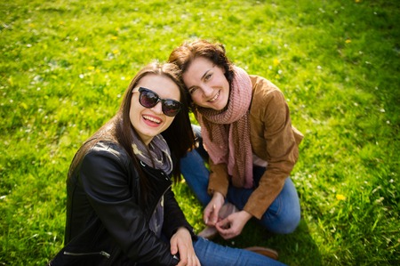 Two young women have a rest on a green lawn. Cute girlfriends joyfully smile. Fine spring day.の写真素材