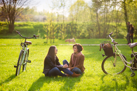 Charming young women sit on a green lawn in the spring park. Their bicycles stand nearby. Girlfriends with enthusiasm about something talk. Fine spring day.の写真素材