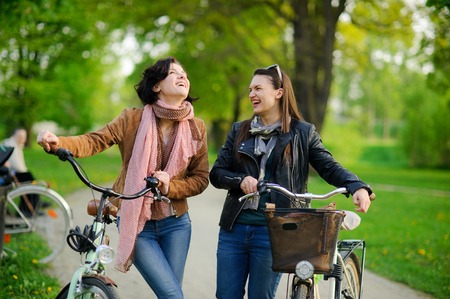 Two charming young women on bicycle walk in the spring park. Girlfriends have stopped and talk. Against the background of fresh spring greensの写真素材