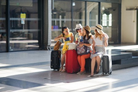 Young tourists waiting for the flight. Girlfriends discuss something. Each girl in a hand has a passport and tickets. Nearby baggage. Summer holiday. Interesting travel.の写真素材