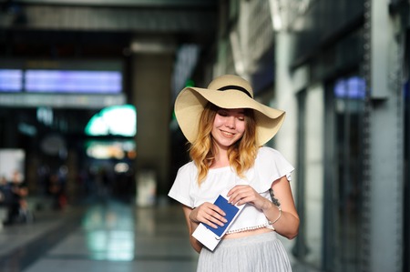 Pretty girl in a wide-brimmed hat with the passport and tickets in a hand. Girl posing. Railway station. Summer holiday.の写真素材
