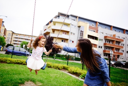Young mother plays in the playground with the little daughter. Woman shakes the baby on a swing. Serene summer day. Good mood.の写真素材