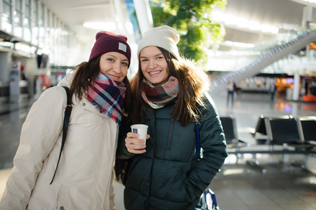 Two young women in the airport waiting room. Girlfriends are happy to pose. They meet someone or expect the flight.の写真素材