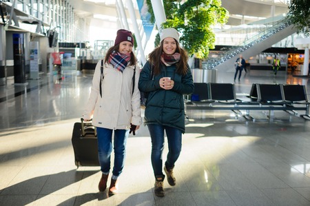 Passengers in the airport waiting room. Young women expect the flight. One of them holds a suitcase. In hands at another - a coffee glass.の写真素材