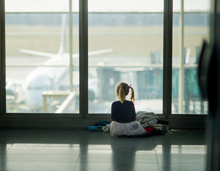 The little girl sits near a big window at the airport. The child looks at big planes on a runway.の写真素材