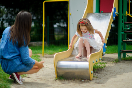 Mom and her little daughter are playing on the playground. Charming baby sliding down a slide at the playground. City court yard. Serene summer day. Good mood.の写真素材