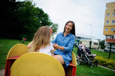 Young mother plays in the playground with the little daughter. Woman and charming girlie turning on the carousel. Serene summer day. Good mood.の写真素材