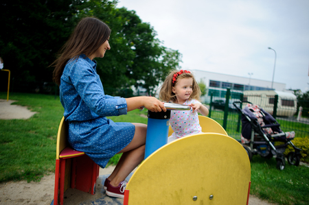 Young mother plays in the playground with the little daughter. Woman and charming girlie turning on the carousel. Serene summer day. Good mood.の写真素材