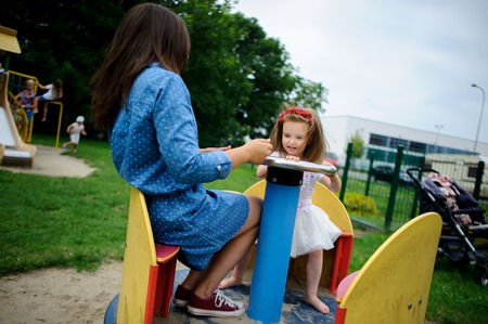 Young mother plays in the playground with the little daughter. Woman and charming girlie turning on the carousel. Serene summer day. Good mood.の写真素材