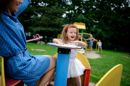 Young mother plays in the playground with the little daughter. Woman and charming girlie turning on the carousel. Serene summer day. Good mood.の写真素材