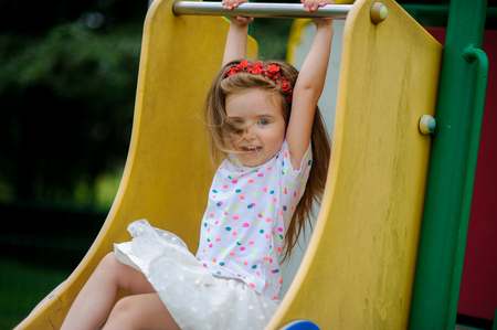 A cute little barefoot girly is playing on the playground. Baby is sitting on the children's slide. On the girl's head is a red wreath.の写真素材