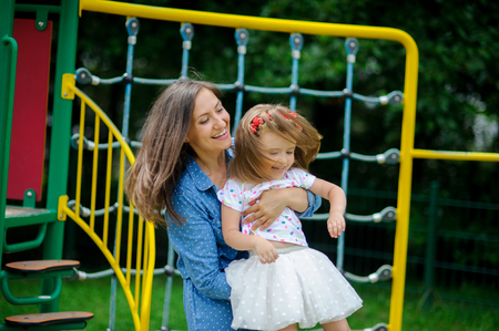 Young mother plays in the playground with the little daughter. Woman holds on hands child. To mother and the daughter it is very cheerful.. Sunny summer day. Good mood.の写真素材
