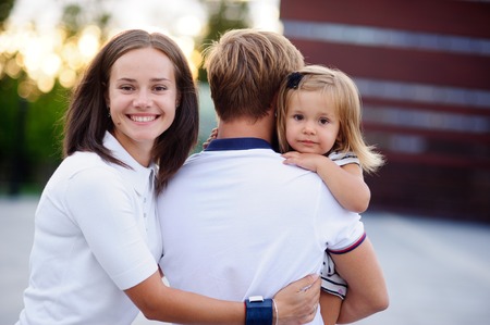 Portrait of a young united family. Father keeps a small daughter in his arms. Woman gently embraces husband and daughter.の写真素材