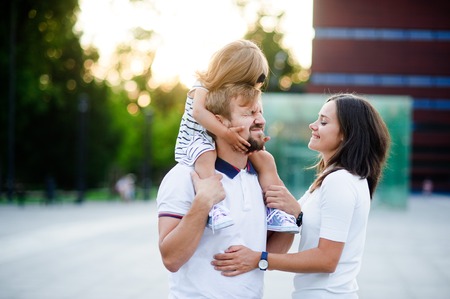 Cute young family on a summer day in the city square. Man holds a small daughter on his shoulders. Woman is hugging her husband. She looks with love and tenderness.の写真素材